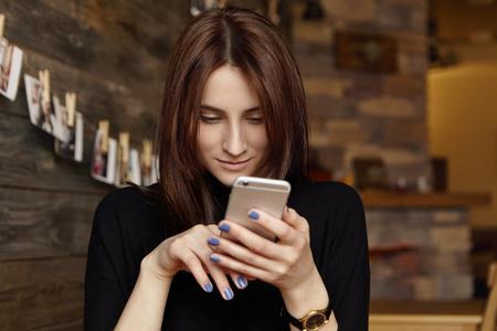 portrait of charming young brunette european lady browsing newsfeed via social networks, liking posts and typing comments, using free high-speed internet connection at coffee shop